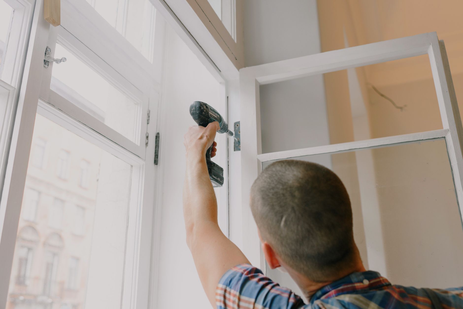 Back view of unrecognizable craftsman using cordless screwdriver and assembling window while screwing metal hinge during reno