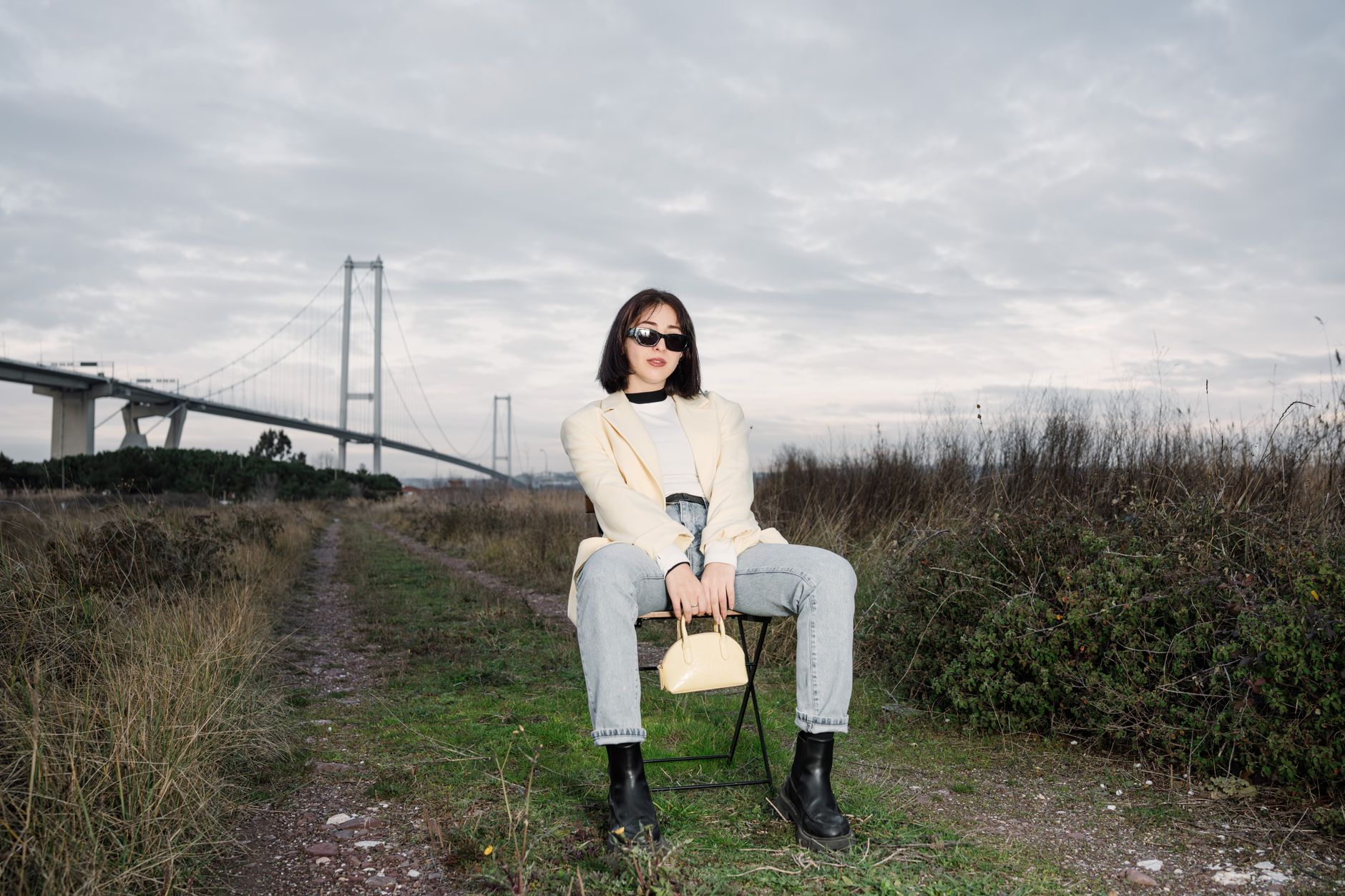 Stylish woman in sunglasses sitting by a bridge, showcasing modern fashion against a natural backdrop. — Finden Sie den beste