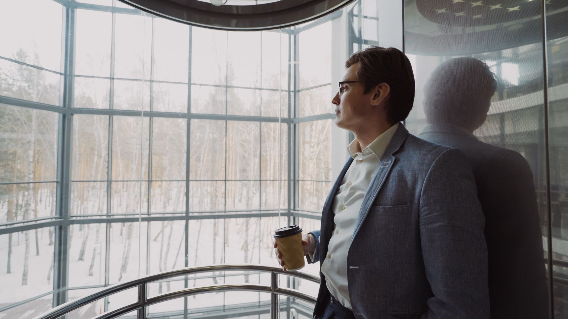 Businessman in blue suit with coffee cup looking out a large window, reflecting on a winter landscape. — Finding the Perfect