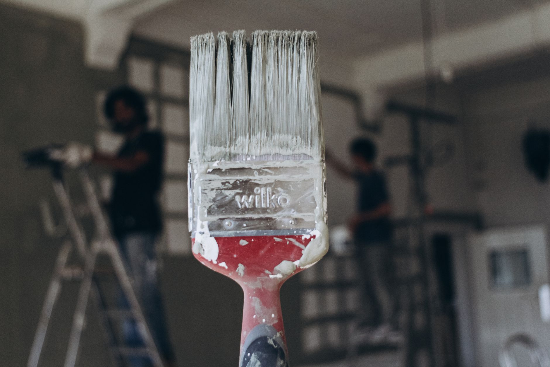 Detailed close-up of a paintbrush with men painting a wall in the blurred background, highlighting renovation work. — he Mode