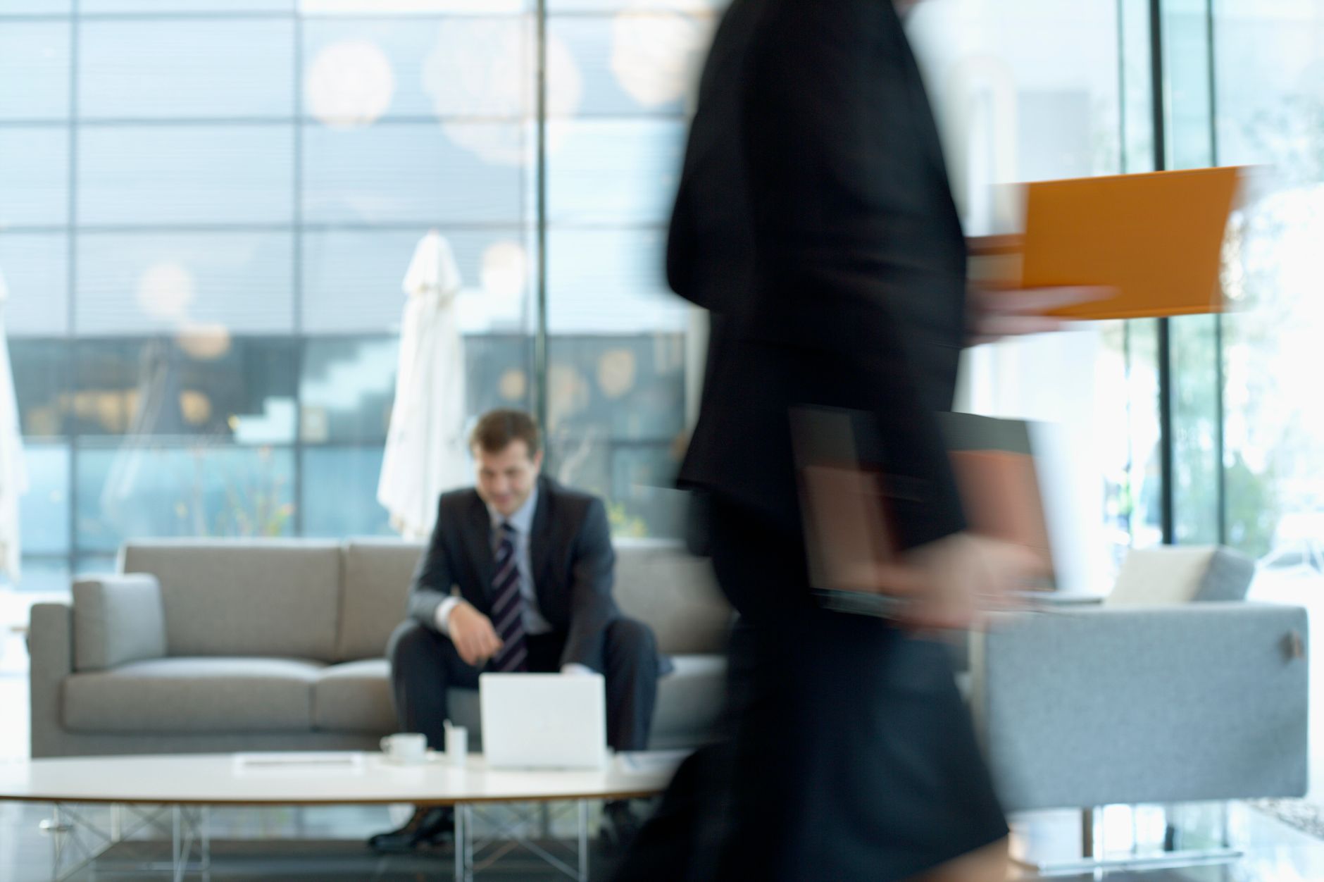 Businessman sitting in modern office lounge using a laptop while colleague walks past, creating dynamic blur. — How to Use n9