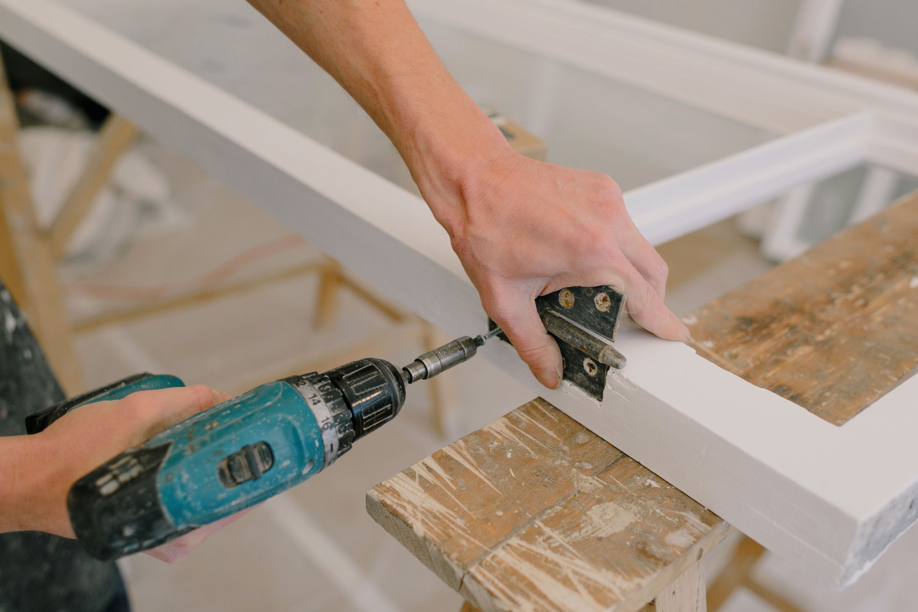 Unrecognizable professional male master using cordless screwdriver to install metal hinge on wooden window in light room duri
