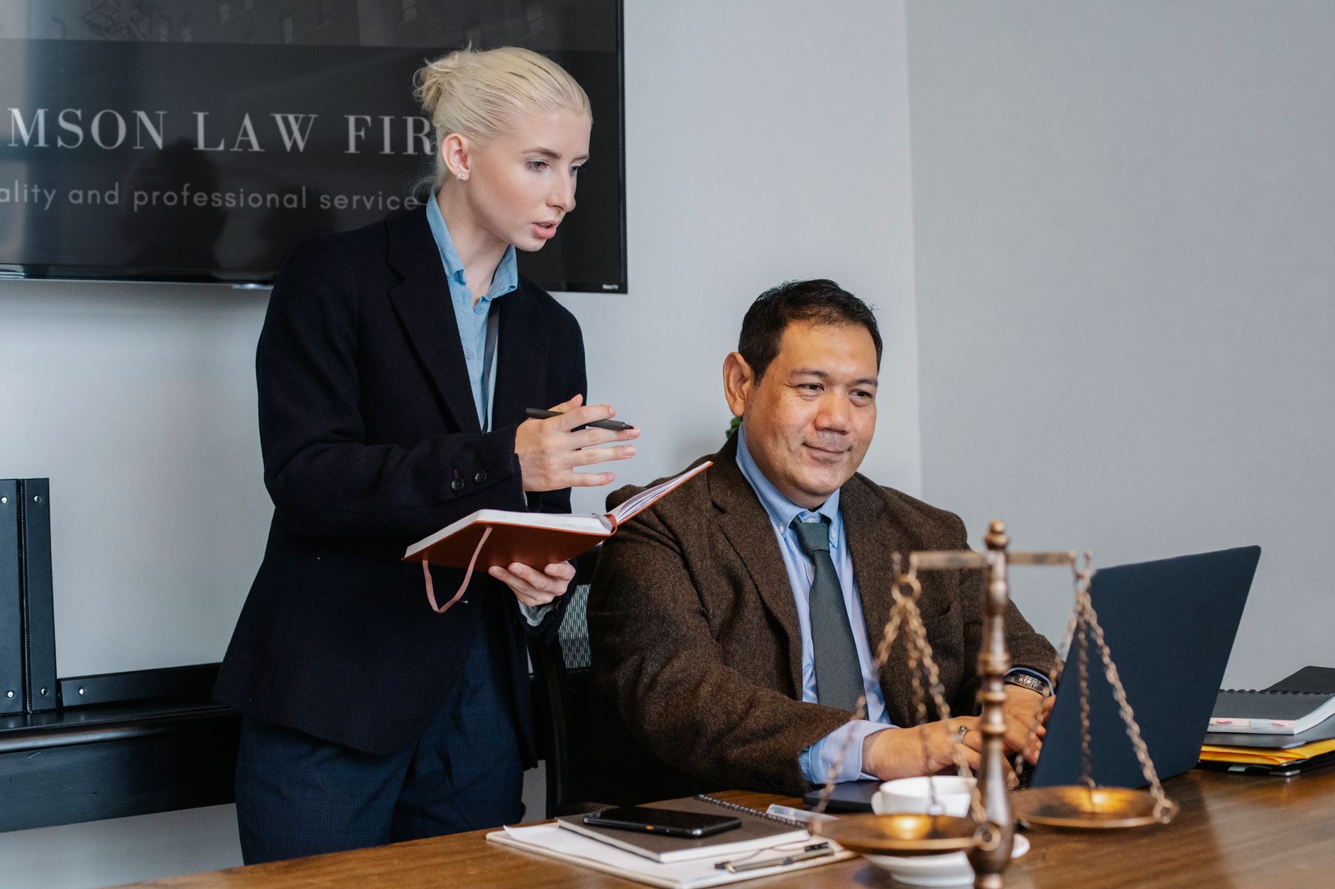 Concentrated young female assistant taking notes in planner while standing at table near middle aged ethnic male colleague ty