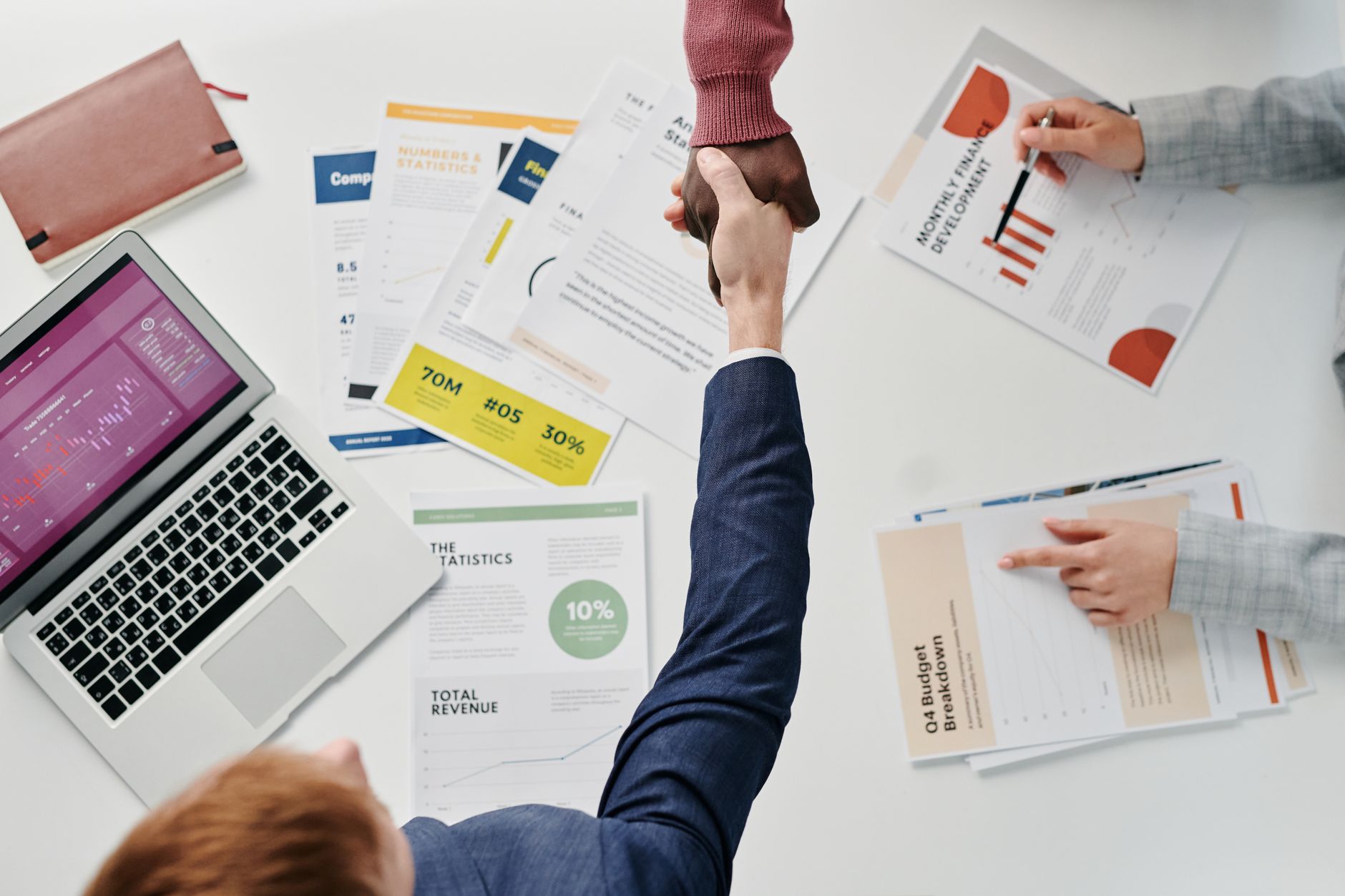 Business professionals engaging in a handshake over a desk filled with financial documents and a laptop. — Jeremy Sch