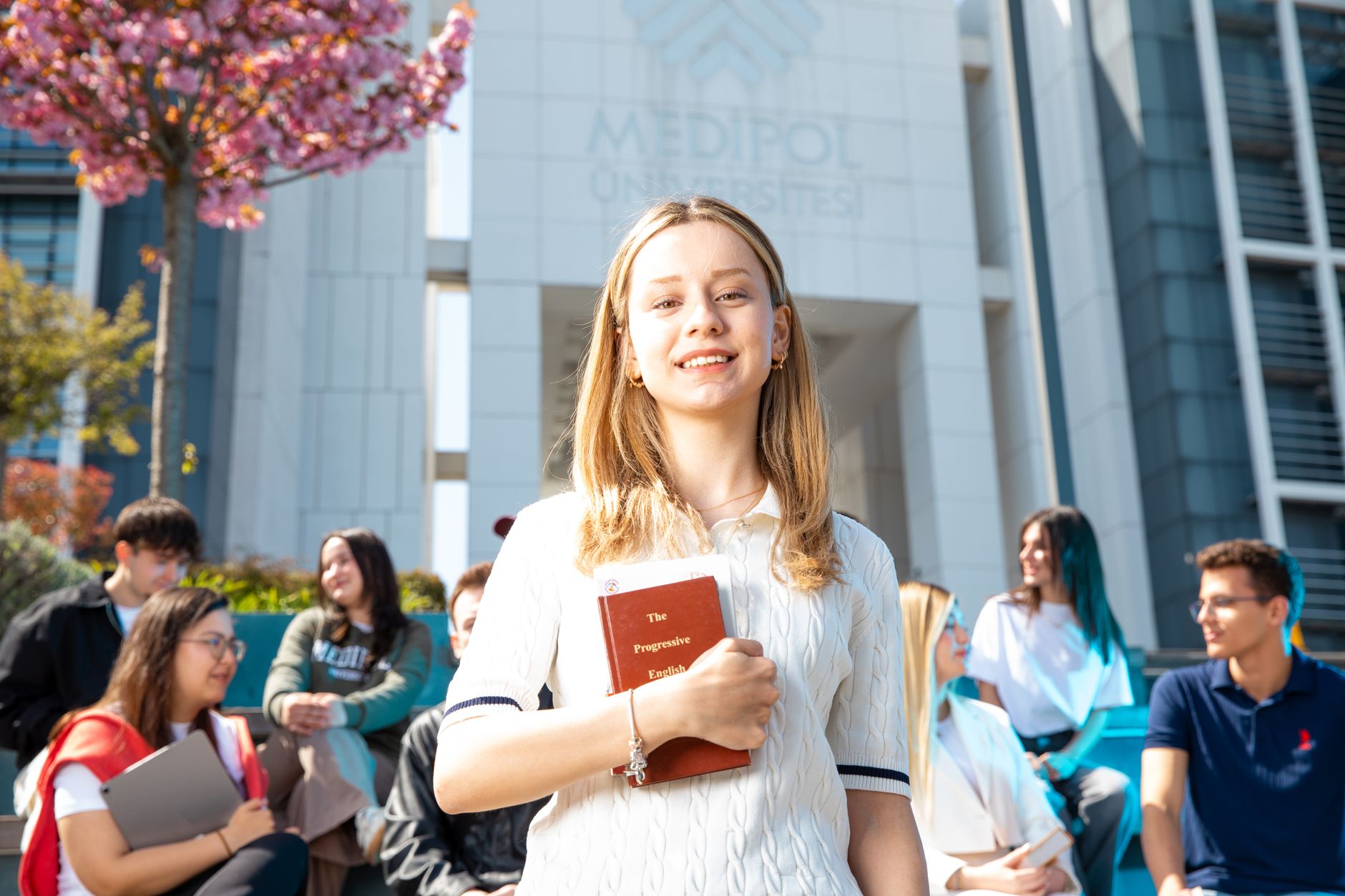 Happy university students socializing on campus, with a focus on a smiling young woman holding a book. — Using Promot