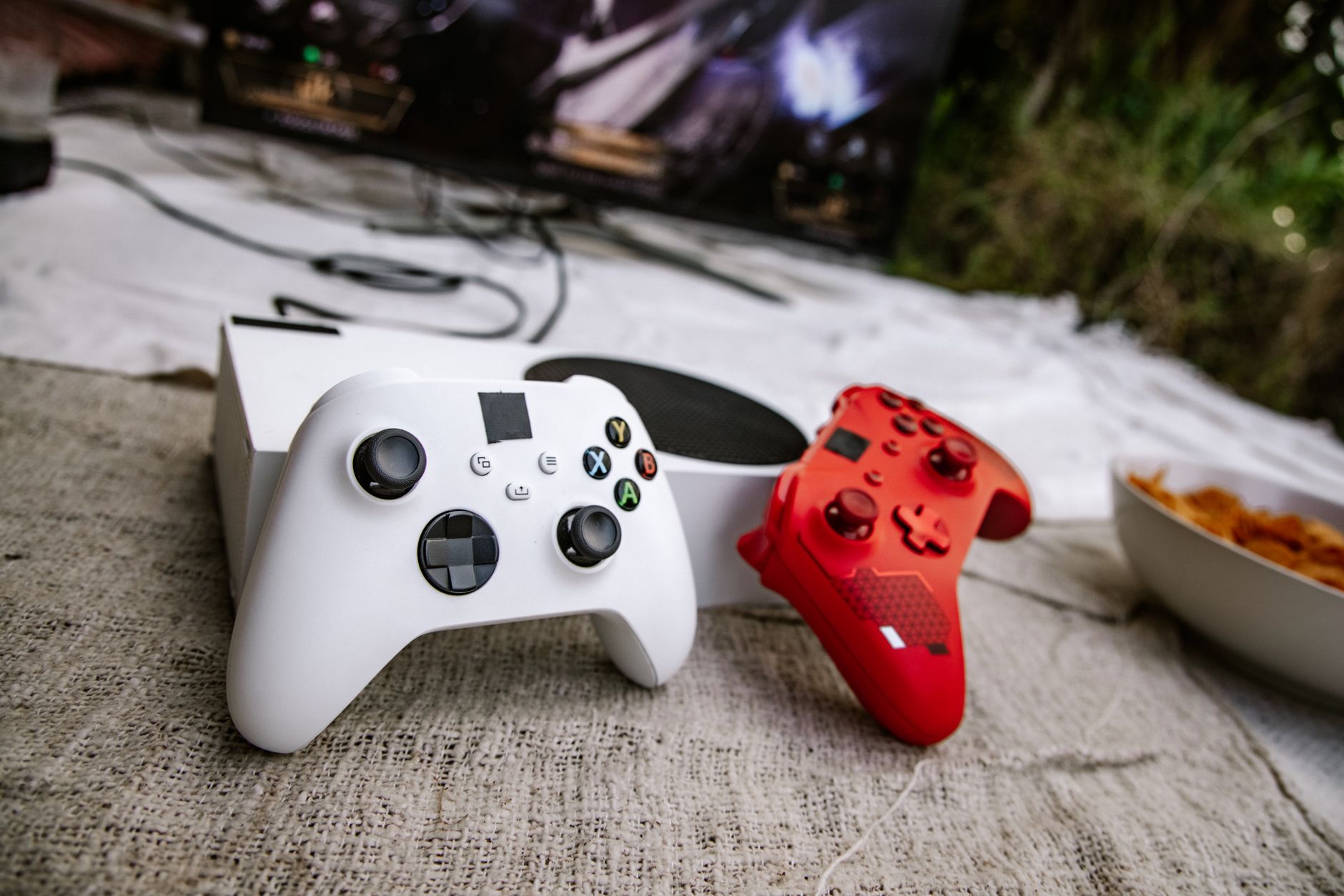 White and red wireless gaming controllers on a textured surface near chips and a TV screen. — Title: Winning Elegance: LSM99